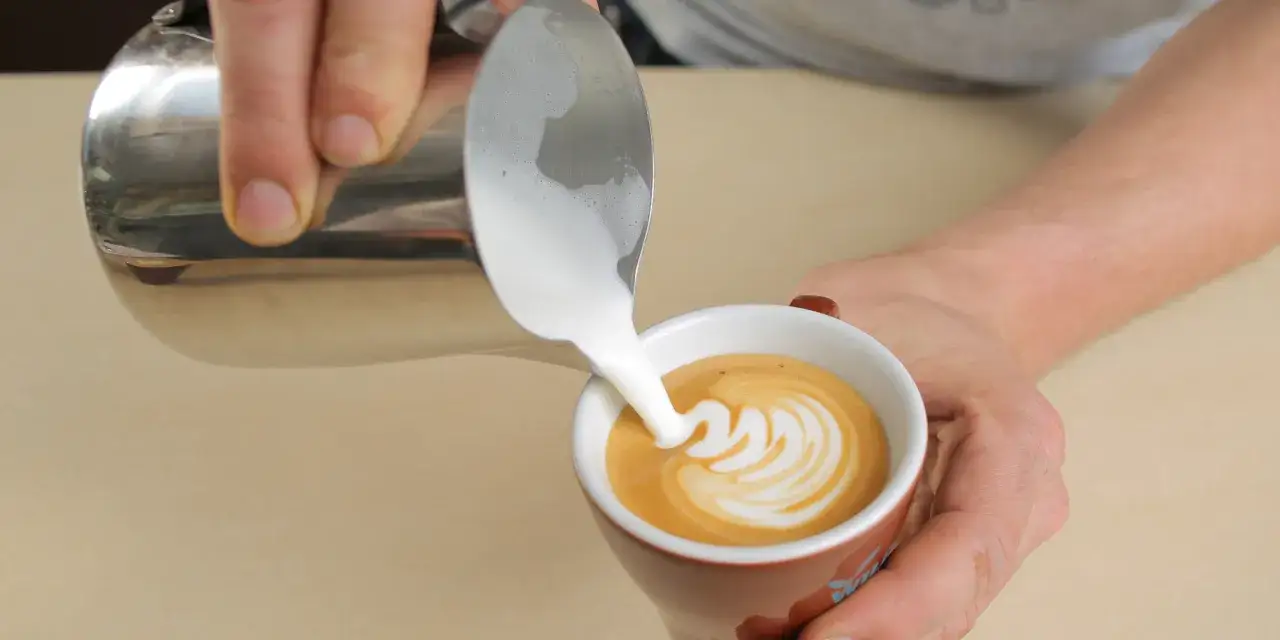 Ręce tworzą latte art, nalewając spienione mleko do kawy. Sztuka spieniania mleka w praktyce.