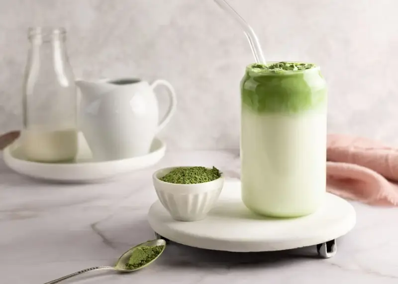 Pyszny matcha latte w szklance, obok miseczka z zielonym proszkiem i łyżeczka.